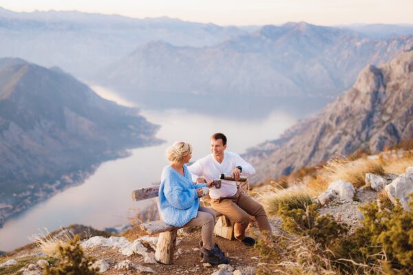 Couple Sits On A Bench And Drinks Tea Against The Backdrop Of Mount Lovcen And The Bay Of Kotor