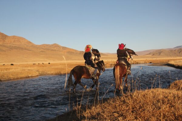 Picture Of Two Horseriders In A River Surrounded By A Deserted Valley With Hills On The Background